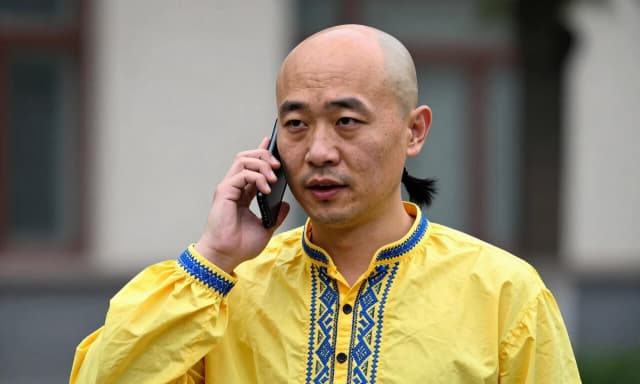 A bald Chinese man is talking on the phone; he is wearing a yellow Ukrainian national shirt with blue embroidery, and a black ponytail hangs to the side from the crown of his head.