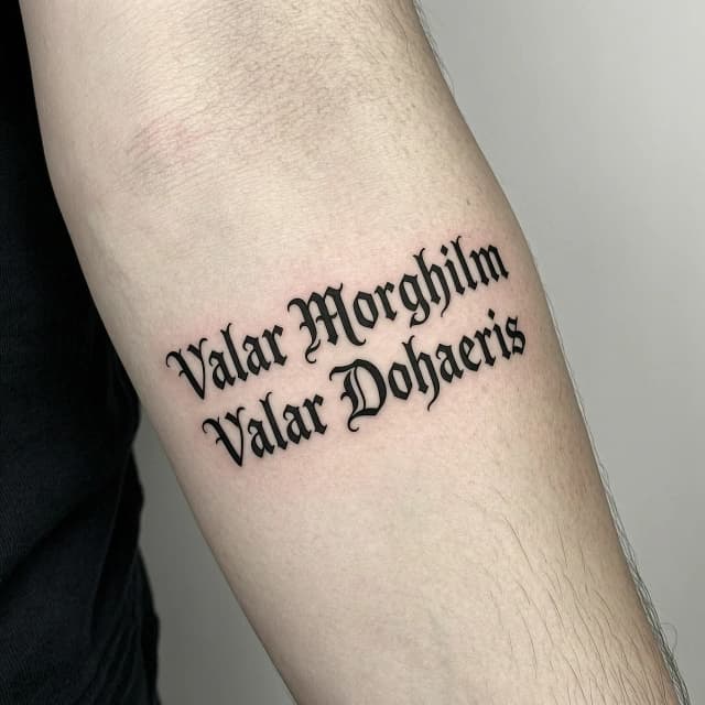 Татуировка на коже предплечья: две строки текста на высоком валирийском «Valar Morghulis» и «Valar Dohaeris». Шрифт строгий, каллиграфический, будто выгравирован на металле. Тонкие чёткие линии, глубокий чёрный цвет, минимум теней. Реалистичный стиль, крупный план, акцент на текстуру кожи и контраст чернил. Высокая детализация.