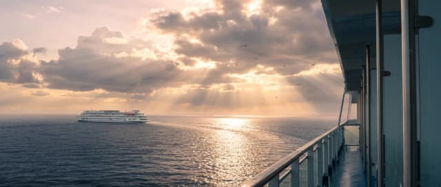 Redraw majestic wide-angle, cinematic landscape shot taken from the elevated deck of a large luxury vessel, looking out over a vast, tranquil ocean during a dramatic sunset. In the mid-ground, a white multi-deck premium cruise ship glides gracefully across the water, leaving a soft, shimmering wake trailing behind it. The sky is filled with heavy, voluminous warm clouds that catch the golden and pale pink hues of the setting sun, creating striking light rays that pierce through the breaks in the cloud cover. The sunlight reflects brilliantly off the rippling surface of the deep blue sea, creating a path of sparkling light that leads toward the horizon. In the foreground, the architectural elements of the viewing deck are visible on the right side, including sleek metallic railings and structural pillars that frame the scene and provide a sense of depth and perspective. A few distant birds are captured in flight, adding a sense of life and scale to the expansive environment. The overall atmosphere is one of serene, melancholic romantic beauty and quiet grandeur, rendered in a realistic photographic style with a soft, dreamlike quality and a palette of deep blues, warm golds, and muted greys.
hyper-realistic Cinematography, and the serene atmosphere of luxury travel.