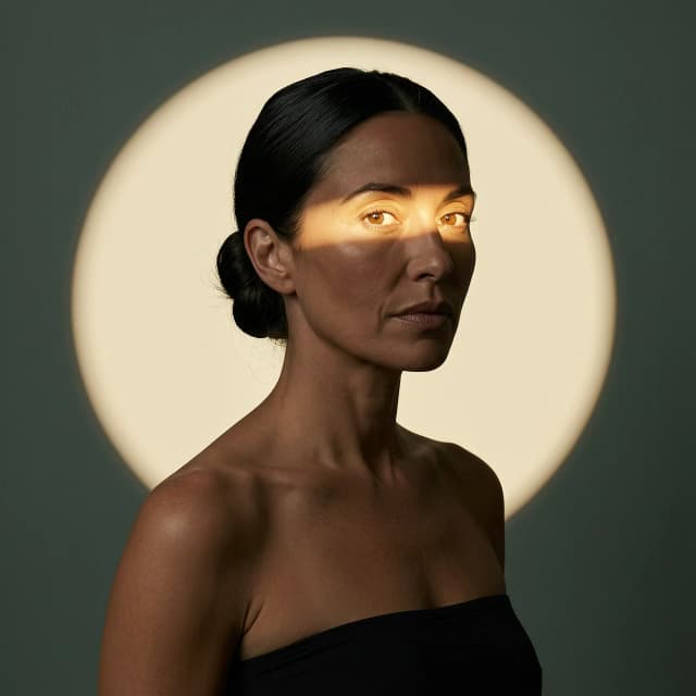 Photorealistic 3:4 portrait of a woman with dark hair in a low bun, black strapless top, dark green-gray background. Large warm cream circular spotlight behind her. A narrow horizontal beam of warm light illuminates only her eyes and upper cheeks, while the rest of her face and body stay in deep natural shadow. Realistic skin texture, natural makeup, high contrast, moody editorial photography, HD.