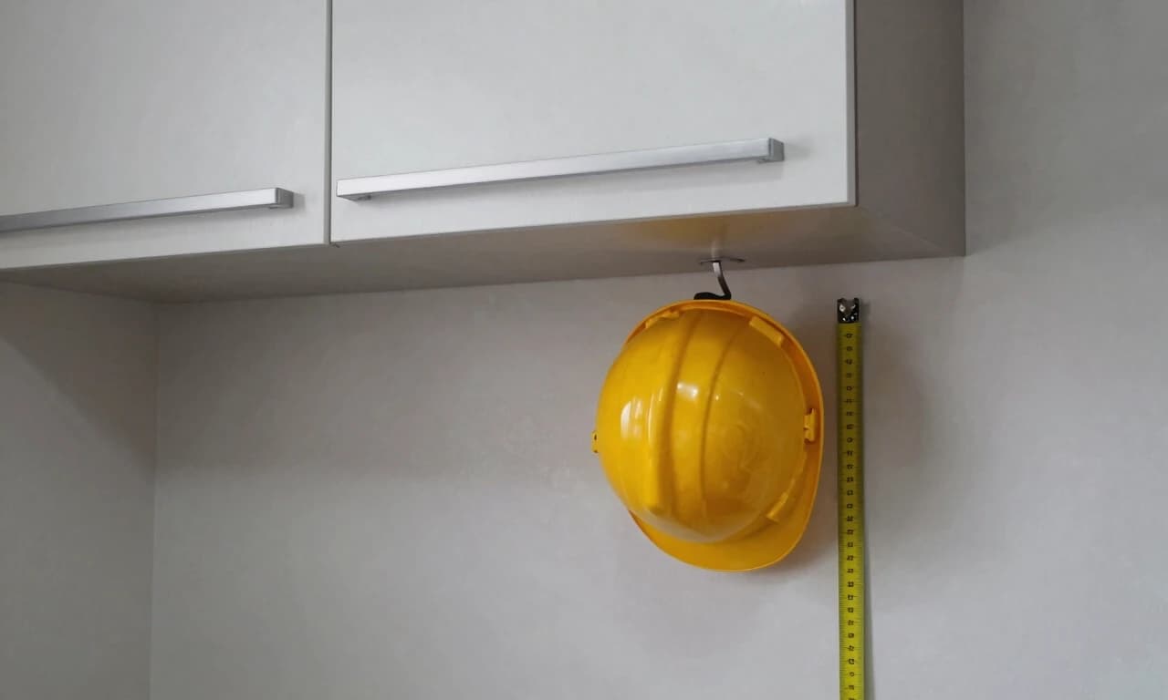 interior photo of a small kitchen with wall-mounted upper cabinets, depth of 35cm, a yellow hard hat hanging from a hook below the cabinet to indicate head clearance issue, measuring scale stuck on cabinet side, tight workspace below, no people, no hands, no text on image, realistic overcast lighting, 8k --ar 16:9 --style raw