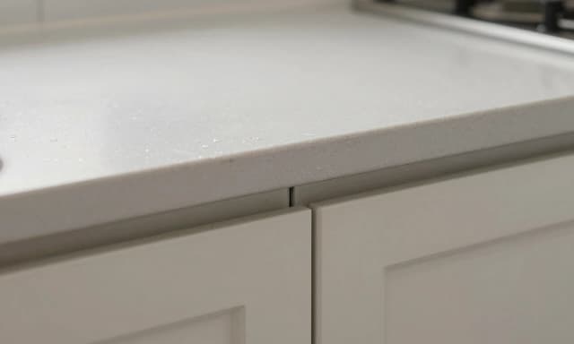 close-up photo of a kitchen countertop edge and cabinet doors, showing a 30mm overhang, water drops on the surface, edge banding slightly swollen at the bottom of doors, no handles visible, realistic wear details, neutral colors, no people, no human hands, no english letters or signs on objects, 8k --ar 16:9 --style raw