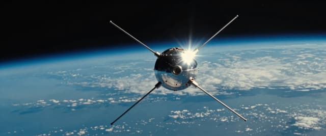 A classic scientific illustration of the Sputnik 1 satellite in orbit high above Earth. The satellite is a polished metal sphere with four long, thin antennas extending in different directions. The image shows the satellite from the side, appearing to be flying to the left. A bright glare of sunlight reflects sharply off the sphere's surface. Below, the arc of Earth is visible, filling the background with layers of blue atmosphere, white clouds, and dark oceans, fading into the blackness of space. The style is vintage sci-fi, with realistic textures and a cinematic composition.