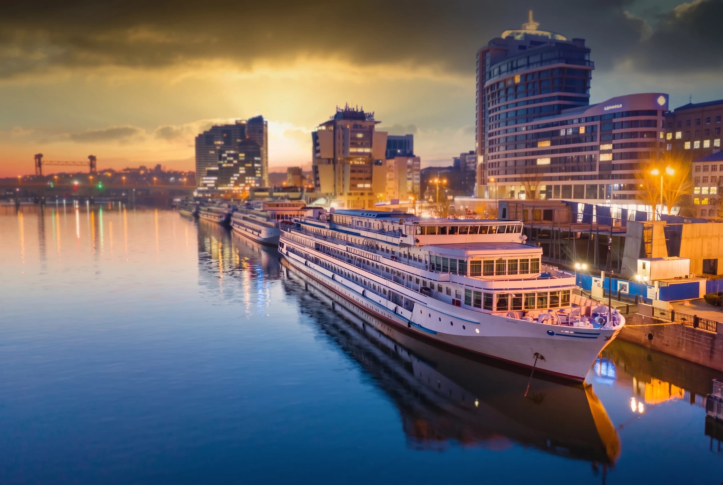 Redraw majestic high-resolution wide-angle cinematic landscape captures a serene riverfront at twilight, dominated by a long, elegant luxury white river cruise ship docked parallel to a stone embankment. The vessel, featuring multiple decks with glowing cabin windows and a sleek, streamlined hull, stretches diagonally through the lower right foreground, its pristine white surface reflecting softly in the calm, glassy water. All attention and visual focus should be directed at the luxury cruise ship. Everything in the surroundings should be either slightly blurred or unobtrusive. It is important to preserve the recognizability and retain exact appearance of the cruise ship. In the background, a modern Russian urban skyline rises against a dramatic sunset, characterized by a mix of high-rise apartment buildings and commercial structures with illuminated windows. The sky is a breathtaking gradient of deep amber, burnt orange, and dusky gold, with heavy, textured clouds catching the last rays of the sun, creating a warm, glowing halo over the city. The lighting is a sophisticated interplay between the natural warmth of the sunset and the artificial cool and gold glows from streetlamps, building windows, and the ship's interior.
