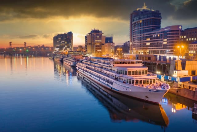Redraw majestic high-resolution wide-angle cinematic landscape captures a serene riverfront at twilight, dominated by a long, elegant luxury white river cruise ship docked parallel to a stone embankment. The vessel, featuring multiple decks with glowing cabin windows and a sleek, streamlined hull, stretches diagonally through the lower right foreground, its pristine white surface reflecting softly in the calm, glassy water. All attention and visual focus should be directed at the luxury cruise ship. Everything in the surroundings should be either slightly blurred or unobtrusive. It is important to preserve the recognizability and retain exact appearance of the cruise ship. In the background, a modern Russian urban skyline rises against a dramatic sunset, characterized by a mix of high-rise apartment buildings and commercial structures with illuminated windows. The sky is a breathtaking gradient of deep amber, burnt orange, and dusky gold, with heavy, textured clouds catching the last rays of the sun, creating a warm, glowing halo over the city. The lighting is a sophisticated interplay between the natural warmth of the sunset and the artificial cool and gold glows from streetlamps, building windows, and the ship's interior.
