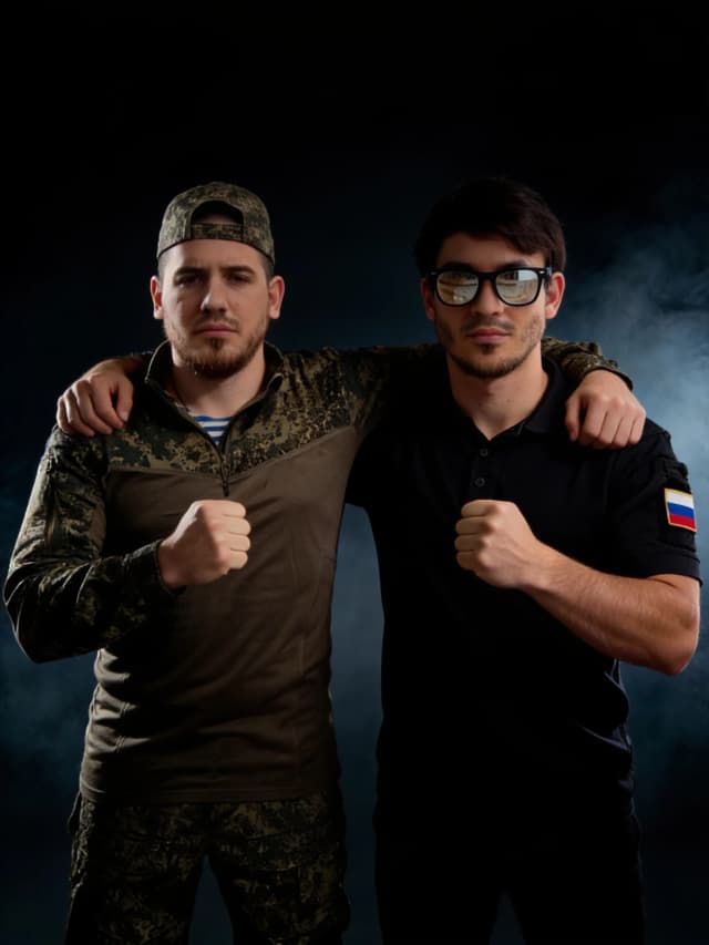 Two people standing side by side in a symmetrical composition, facing the camera with a direct and intense gaze. They are embracing each other by placing one arm over each other's shoulders in a gesture of solidarity. Simultaneously, each person holds a tightly clenched fist in front of their chest with their other hand. Cinematic portrait photography, dramatic chiaroscuro lighting with strong highlights and deep shadows to emphasize muscle tension and facial expressions. Hyper-realistic textures, detailed skin pores, and expressive eyes. The background is a dark, moody, atmospheric studio setting with subtle volumetric smoke. High contrast, epic and powerful mood, 8k resolution, shot on 85mm lens, sharp focus, masterpiece quality.