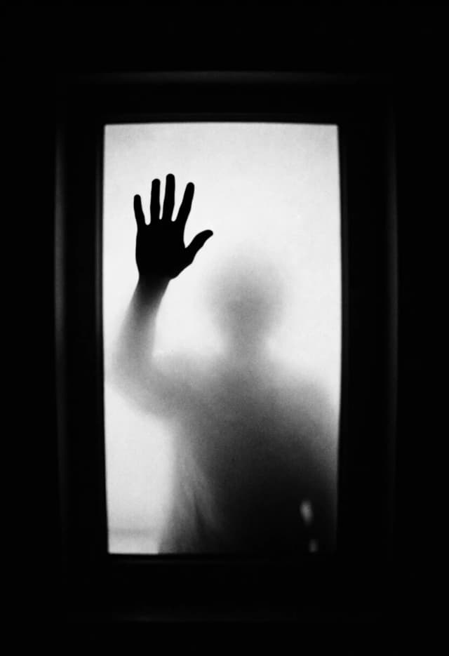 Нарисуй вертикально ориентированную композицию в стиле высококонтрастной чёрно‑белой фотографии. В центре — прямоугольный проём (окно, дверь или портал), заполненный плотным, слегка зернистым светом. Текстура фона должна имитировать старую фотоплёнку или мелкозернистую бумагу: добавь равномерный шум, едва заметные крапинки и лёгкую неоднородность тона, чтобы усилить ощущение винтажности и отчуждённости.
В верхней левой части светового проёма размести чёткий чёрный силуэт человеческой ладони — пальцы слегка разведены, контур резкий, без градиентов, будто вырезан из абсолютной тьмы. Ладонь должна выглядеть как застывший жест: не прижатая к поверхности, а будто парящая в миллиметре от неё — это создаст напряжение и ощущение барьера.
Под ладонью и ниже по центру проёма добавь размытый, призрачный силуэт человеческой фигуры — не детализированный, а как лёгкое затемнение, дымка или тень: полупрозрачный, с плавным переходом от более тёмного пятна к едва уловимому контуру. Он не должен отвлекать от руки, но обязан считываться как присутствие.
Проработай контраст: края светового проёма оставь идеально чёрными, без полутонов, чтобы они выглядели как глубокая пустота. Сам свет внутри проёма сделай неоднородным: в верхней части — ярче, почти белым, к центру и низу — мягко темнеющим, с лёгкими градиентами и едва заметными круговыми ореолами, будто от старого объектива.
Сохрани минимализм: никаких лишних деталей, узоров или отвлекающих элементов. Вся драматургия строится на контрасте света и тьмы, зернистой фактуре и двух ключевых образах — руке и призрачном силуэте.
Палитра: строго монохромная (глубокий чёрный, оттенки серого, чистый белый). Стиль: нуарная фотография, зернистость, высокий контраст, кинематографичная композиция, 8K, акцент на атмосферу отчуждения и ожидания.
