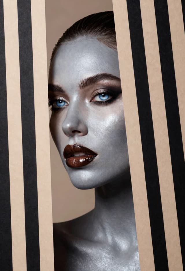 Create an image of a woman partially hidden by a series of vertical black and beige striped patterns. The woman is strikingly beautiful. The shade of a woman's skin is a smooth, cool shade of light gray, which gives her a metallic look. It has striking features: high cheekbones, a pointed nose and full lips covered with a shiny, deep brown tint. Her eyes are captivating icy blue, surrounded by dark eyelashes and arched eyebrows, which accentuate her intense gaze. The striped patterns create visual distortions, seamlessly merging into her contours, while revealing only parts of her features, strengthening the sense of depth and intrigue. The background consists of alternating black and beige stripes that continue the pattern, creating a strikingly modern and artistic composition. The light source casts subtle shadows, adding dimension to her profile, while emphasising the shiny finish of her lips and the smooth texture of her skin. The general tone is avant-garde, combining beauty with abstract art, emphasising the contrast between what is organic and what is geometric.