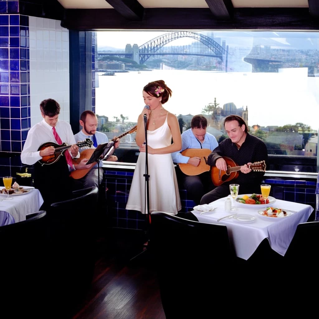 The image shows a group of people in a restaurant with a beautiful view of the city. The restaurant has blue and white tiles on the walls and a large window that offers a panoramic view of a bridge and buildings. In the center of the image, there is a woman in a white dress with a flower in her hair, singing into a microphone. To her left, there are two men playing mandolins, one playing a mandolin and the other playing a guitar. To the right, two men are playing guitars, and there are plates of food and drinks on the table in front of them. The overall atmosphere of the restaurant is romantic and lively.