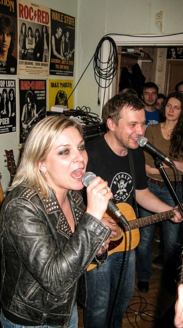 Hyper-realistic candid snapshot from the mid-2000s, captured on a digital compact camera with slight motion blur and authentic film grain. A medium shot of a punk-rock female singer performing at a small, intimate underground apartment concert (kvartirnik), standing side-by-side with Alexander Pushnoy. The woman has her original facial features preserved, styled with messy punk hair, heavy smudged eyeliner, and a studded leather jacket. Alexander Pushnoy is depicted with his authentic likeness, looking natural and engaged in the atmosphere. The composition is slightly unpolished and spontaneous, like a real fan photo. Lighting is harsh and uneven, coming from a direct camera flash, creating deep shadows against the cluttered background of a cozy, dimly lit Soviet-style apartment with posters on the walls, cables, and musical instruments. High fidelity to human proportions, realistic skin textures with natural imperfections, and authentic color grading typical of early 2000s digital photography. No artificial smoothing, no airbrushing, looks like a genuine, unedited archival photo.